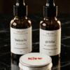 IMANY set prirodne kozmetike na beloj pozadini. Set sadrži dve tamne staklene bočice sa pipetom: "geisha" ulje za regeneraciju kose (30ml) i "bakuchi" anti-age ulje za lice (30ml), kao i jednu malu okruglu srebrnu limenku "NEŽAN" balzama za usne sa crvenim srcem na etiketi.