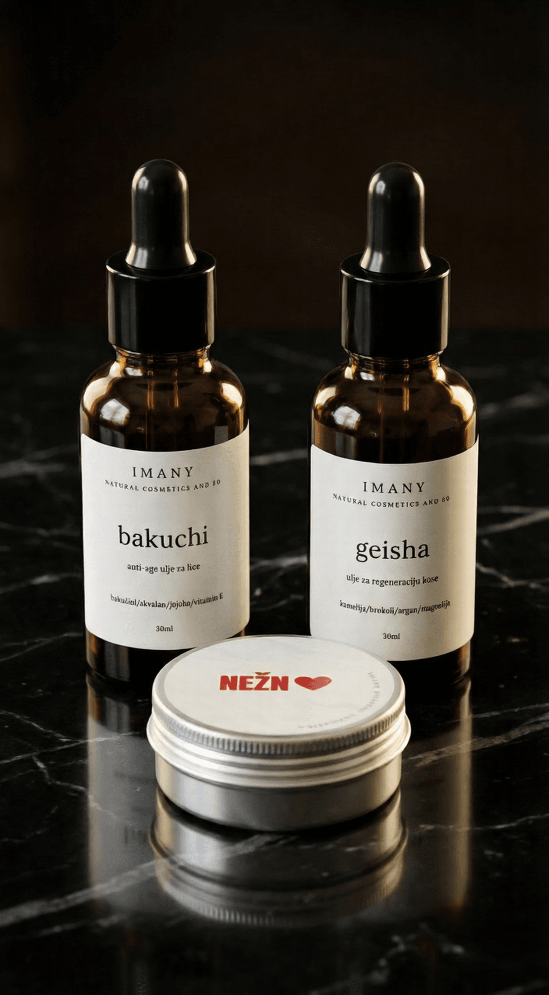 IMANY set prirodne kozmetike na beloj pozadini. Set sadrži dve tamne staklene bočice sa pipetom: "geisha" ulje za regeneraciju kose (30ml) i "bakuchi" anti-age ulje za lice (30ml), kao i jednu malu okruglu srebrnu limenku "NEŽAN" balzama za usne sa crvenim srcem na etiketi.