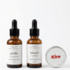 IMANY set prirodne kozmetike na beloj pozadini. Set sadrži dve tamne staklene bočice sa pipetom: "geisha" ulje za regeneraciju kose (30ml) i "bakuchi" anti-age ulje za lice (30ml), kao i jednu malu okruglu srebrnu limenku "NEŽAN" balzama za usne sa crvenim srcem na etiketi.